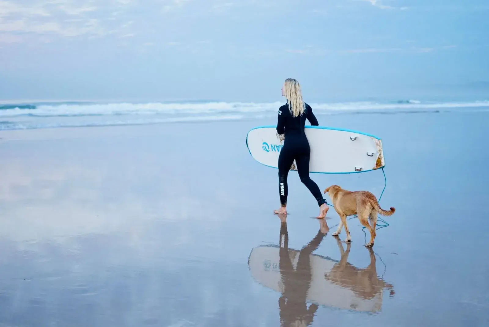 Surfer carrying a white board along the water’s edge beside a dog